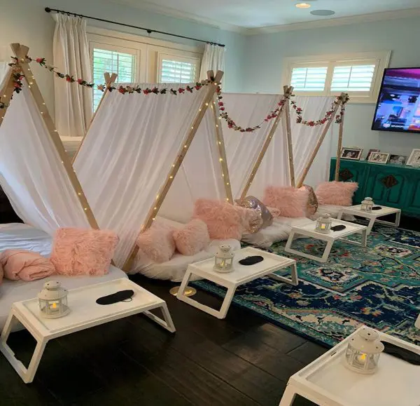 A cozy indoor setup with teepee tents adorned with fairy lights and floral garlands, cushioned seating, small tables with lanterns, and a TV on the wall in a room with a patterned rug—a perfect kids party rental for a magical slime party.