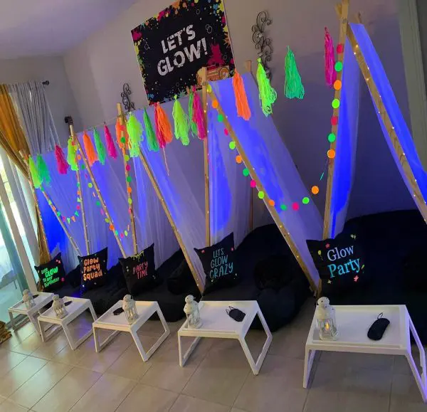 Indoor setup for a glow-themed slime party featuring teepees with neon decorations, pillows with glow-related text, low tables with lanterns, all under a "Let's Glow!" banner. Perfect for a kids party rental experience!