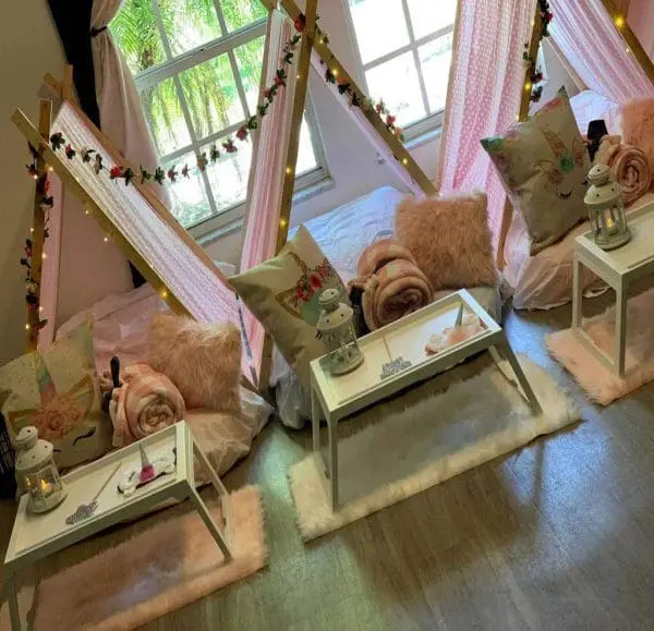 Three indoor teepee tents with pink decor, two pillows each, surrounding white tables. Blankets and lanterns are placed on the tables, perfect for a cozy slumber party. The window in the background shows greenery outside. Ideal for a charming party rental setup.