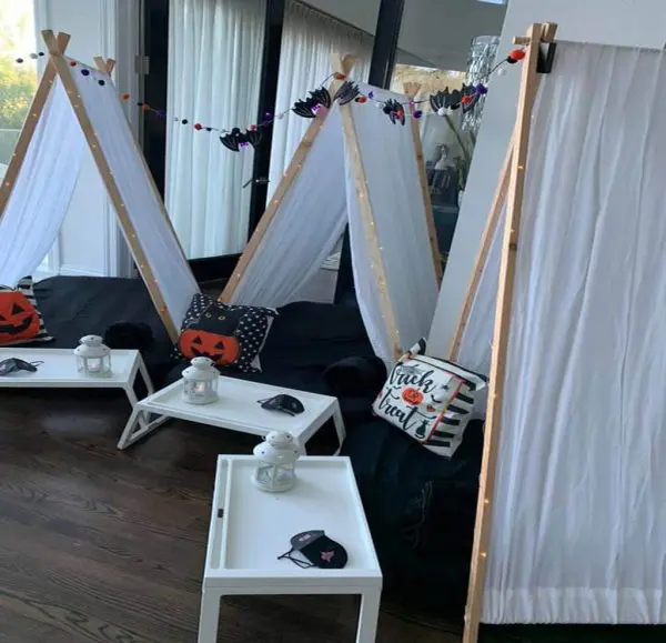 Two white fabric tents decorated with Halloween-themed pillows and garlands are set up in a room, perfect for a kids party rental. Small tables with lanterns and eye masks are placed in front of the tents, adding to the festive atmosphere.