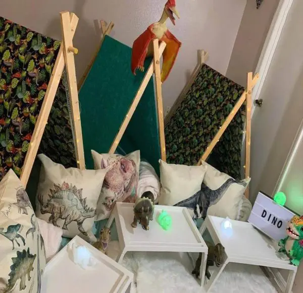 Three small wooden teepees with green and dinosaur-patterned fabric, surrounded by dinosaur-themed pillows, toy dinosaurs, white tables, and soft lighting. A toy Pteranodon is perched on one of the tents. Perfect for a fun and memorable slime party or any themed party rental occasion.