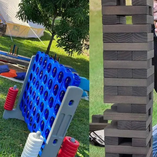 Two oversized outdoor games from our XL Outdoor Games Addon: a giant Connect Four with red and white discs, and a large Jenga tower on a grassy lawn. A tent and trees in the background make it perfect for any fun-filled event.