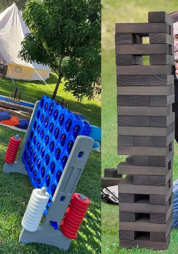 Two oversized outdoor games from our XL Outdoor Games Addon: a giant Connect Four with red and white discs, and a large Jenga tower on a grassy lawn. A tent and trees in the background make it perfect for any fun-filled event.