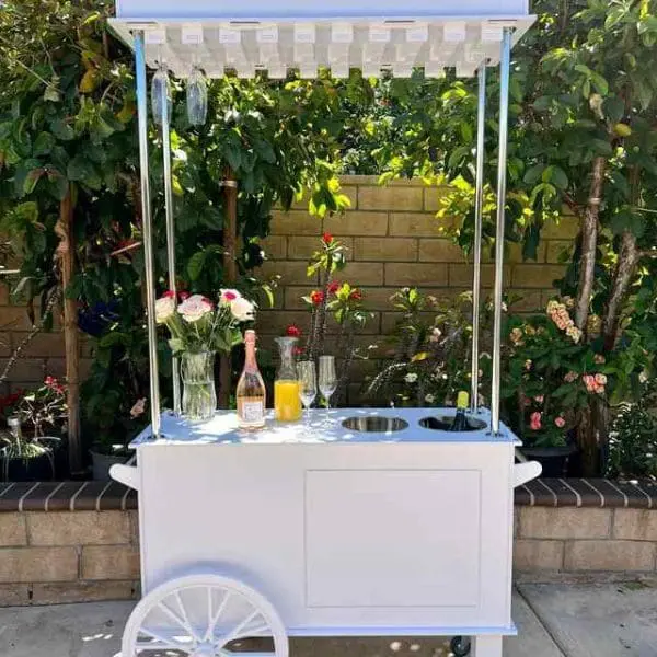 The XL Outdoor Games Addon includes a white cart with a pergola roof, set in a picturesque garden. Decorated with vases of flowers and accessorized with a bottle of champagne, two glasses, a pitcher of orange juice, and another pitcher, this setup is ideal as a party rental backdrop. The scene is further enhanced by lush green foliage and an inviting brick wall in the background.