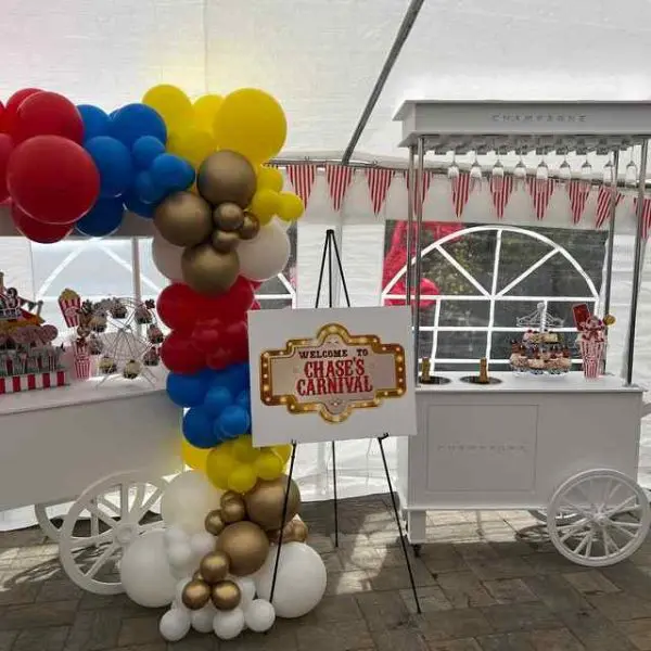 Carnival-themed party setup featuring a colorful balloon display, candy cart, and a sign reading "Welcome to Chase's Carnival," all complemented by the XL Outdoor Games Addon. A white tent in the background is provided by our party rental service.