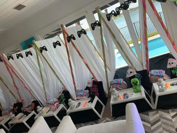 Four indoor teepees are set up in front of large windows overlooking a pool and lake. Each tent has a small table with blankets, plush toys, and lanterns, along with gamer-themed decorations, ready for a cozy party service that blends comfort with fun.