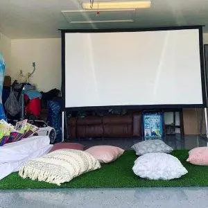 A garage converted into a home theater with a large projector screen, floor pillows, and a piece of artificial turf makes an ideal kids party rental. Various household items and toys are stored in the background, setting the perfect scene for an unforgettable slumber party experience.