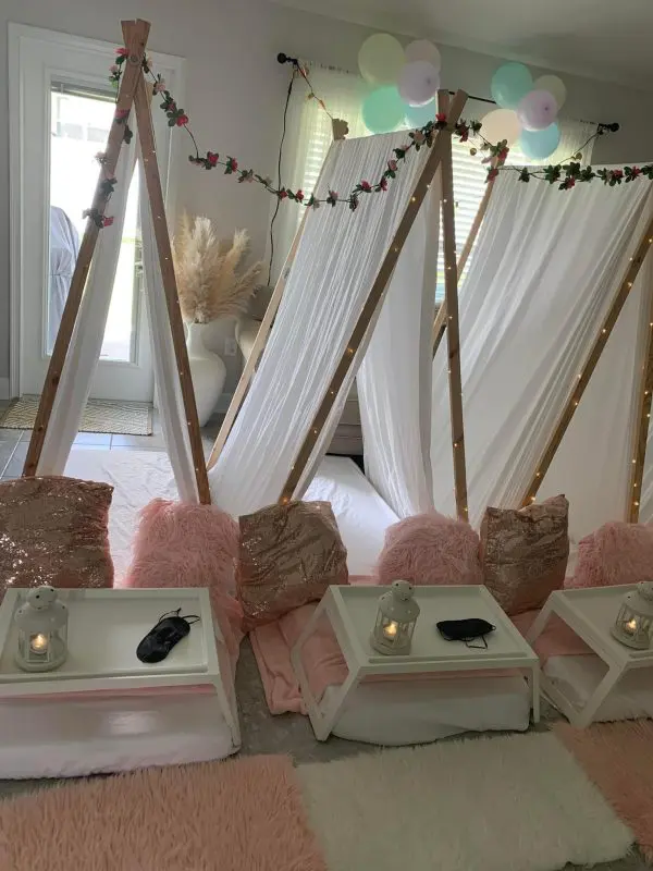 Three indoor teepee setups with white fabric, fairy lights, and floral garlands. Each tent has a small tray with a candle and eye mask on a fluffy rug. Pink pillows and blankets are placed in front. Perfect for your next event, this party rental service ensures a magical experience.