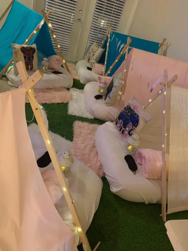 A room with five small indoor teepees, each featuring pillows, blankets, sleeping masks, and small lanterns, all set on a grass-like carpet with a cozy ambiance—perfect for a kids party rental.