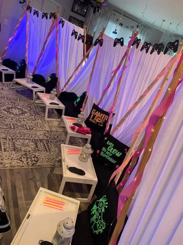 A room set up for a gaming-themed slumber party with white tent-like structures, black game controller decorations, lanterns, and party favor bags. Neon sticks are placed on small tables near the tents, creating an exciting atmosphere perfect for a kids party rental.