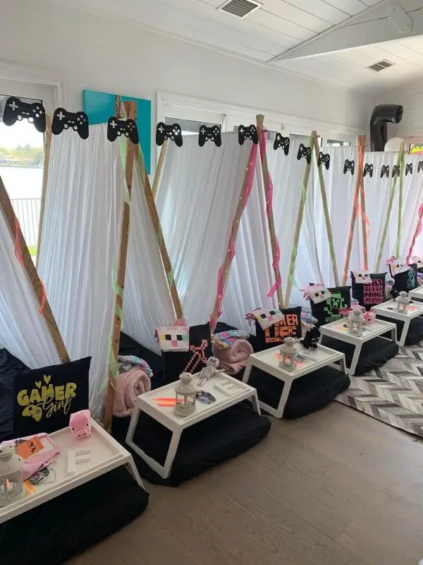 Indoor tent setup with gaming-themed decorations, including black and pink pillows, gamer-themed pillowcases, and controller cutouts above each tent. Perfect for a kids party rental, small trays with snacks and drinks are in front to keep everyone fueled for fun.