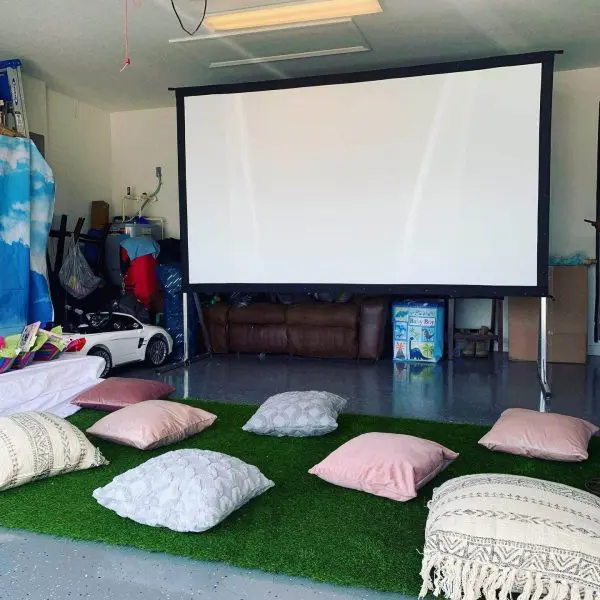 A garage set up for a home theater with a projector screen, a couch, pillows on faux grass for seating, and various items stored in the background—perfect for hosting a cozy slime party.