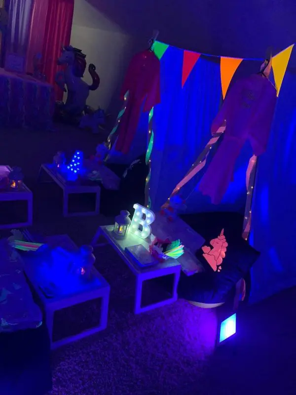 A dimly lit room features glowing decorations, a teepee setup with colorful pennants, illuminated letters, and small tables with various glow-in-the-dark items. A unicorn decoration is also visible, making it perfect for a unique party rental experience.