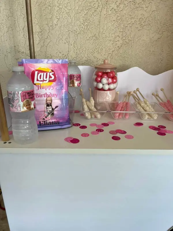 A decorated table for a kids party rental features a birthday Lay's chips bag, water bottles, gumballs in a jar, and candy sticks. A festive touch is added to the table's surface with pink and white confetti scattered around. The setup is completed with a Large Greenery Photo Backdrop featuring a vibrant neon sign.