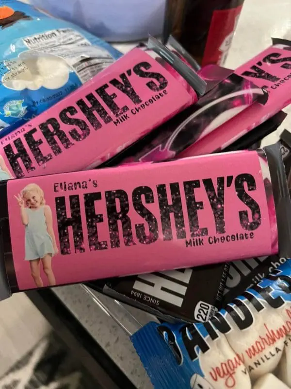 A vibrant setting for a lively slumber party is created with pink-wrapped Hershey's milk chocolate bars, each adorned with "Eliana's" printed above the brand name, scattered across a cluttered surface in front of a Large Greenery Photo Backdrop highlighted by an eye-catching neon sign.