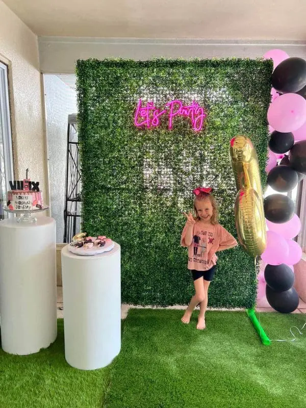 A young girl stands in front of the Large Greenery Photo Backdrop with Neon Sign that says "Let's Party," next to a gold balloon and a display of treats. Black and pink balloons float nearby, creating the perfect scene for an unforgettable slumber party with teepees available for rental.