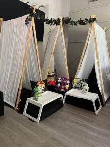 Three indoor tent setups with white fabric, wooden frames, and string lights create a magical slumber party setting. The area includes two small white tables adorned with themed plush toys and decorative pillows on black cushions. Perfect for a whimsical night, this party service ensures unforgettable memories.