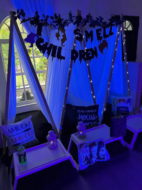 Indoor sleepover setup with decorated teepees, cushions, and themed signs under blue lighting. Signs include "Amuck! Amuck! Amuck!" and "I Smell Children." Small lanterns adorn the scene. Perfect for a slumber party, this unique experience is available through our party rental service.