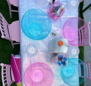 A charming table set with holographic plates, craft supplies, and a container of milk is surrounded by pink chairs on a grass surface—a perfect setup provided by our party rental service.