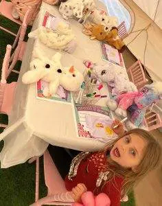 A child sits at a table with stuffed animals, adoption certificates, and markers laid out on the table. The scene appears to be part of a slumber party-themed activity, complete with cozy teepees in the background for an enchanting night of fun.