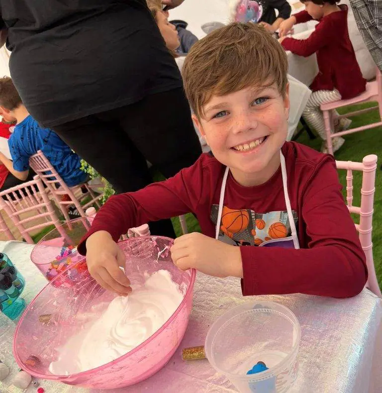 A smiling child in a red shirt plays with a bowl of fluffy white material at a table, surrounded by other children engaging in activities, making for a perfect party rental setting.