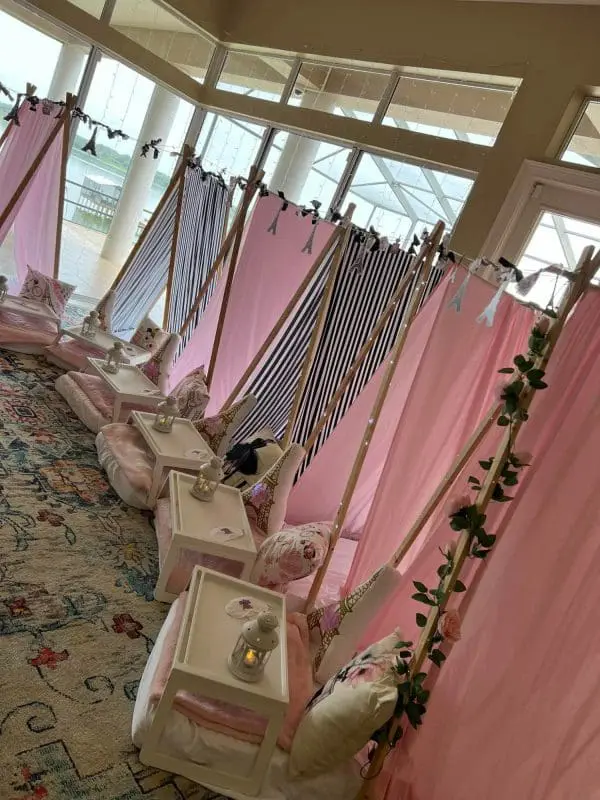 A row of indoor pink and black striped teepees with white cushions and small tables set up for a party rental event. Each table holds a small lantern. The setting overlooks a bright outdoor area through large windows, making it perfect for a memorable party service.