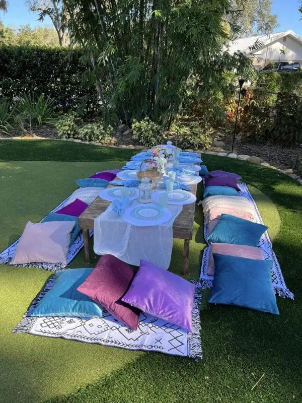 An outdoor dining setup on a grass lawn features a low wooden table with white tableware, a table runner, and a floral centerpiece. Colorful cushions and patterned rugs surround the table for seating, creating the perfect ambiance for an elegant party service.