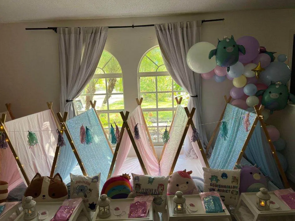 A room setup for a children's sleepover features small tents, colorful decorative pillows, and fairy lights. Perfect for a slumber party, the space is adorned with balloons and curtains framing a large window in the background. Consider this charming scene for your next kids' party rental.