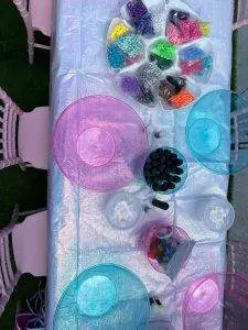 A table with an iridescent tablecloth is set up for a slime party, featuring various colorful beads, two large empty bowls, small containers, and chairs around it, ready for a fun craft activity.