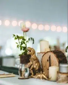 A decorated table centerpiece featuring a gold dinosaur figurine, a small water glass, a vase with a pink flower, candle holders, and a round wooden piece with the number one—perfect for any kids party rental or party service.