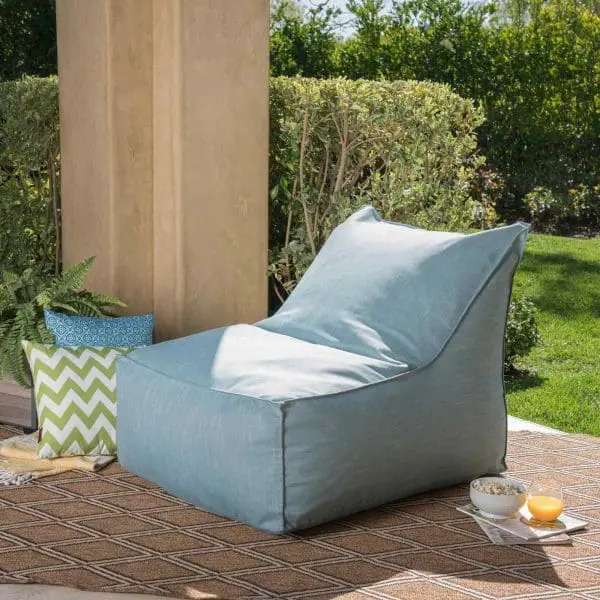 A light blue outdoor lounge chair, perfect for a kids' party rental, is placed on a patterned rug next to a pillar, with green bushes in the background. There are various pillows and a small bowl on the ground beside the chair.