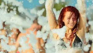 Woman smiling in a black bikini top, enjoying a foam party outdoors, with party rental equipment and other people covered in foam in the background.