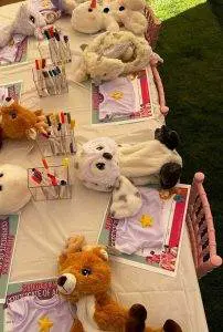 Plush toys and certificates are arranged on a table with activity sheets, colored markers, and small containers, perfect for a cozy slumber party activity session. The setup is designed to entertain children with crafts or activities under teepees or during a slime party.