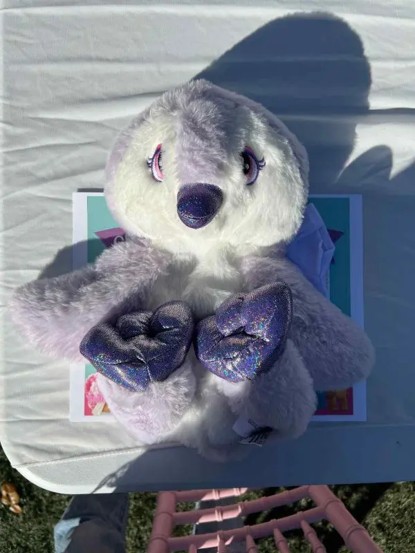 A stuffed animal toy with purple and white fur, glittery bow, and closed eyes lies on a white table in sunlight, ready for the slumber party.