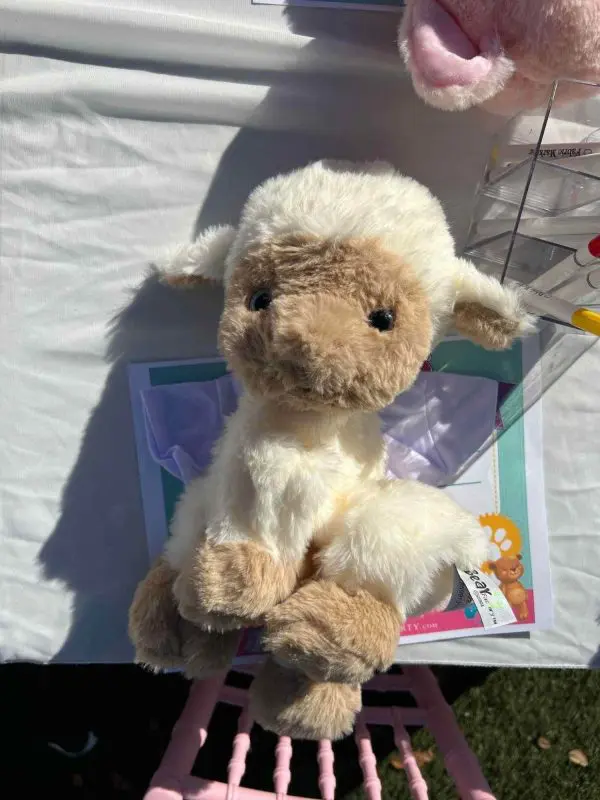 A plush lamb toy with white fur and light brown face, ears, and hooves is placed on a table, perfect for a slumber party. Some plastic packaging can be seen in the background.