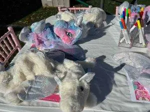 Stuffed unicorns with sequined wings displayed on a table at a Build-A-Bear party, accompanied by colorful markers and small pink chairs.