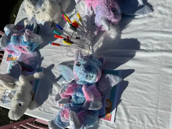 Plush unicorn toys displayed for sale on a table with colorful fabric pens beside them at a bear party, outdoors in bright sunlight.