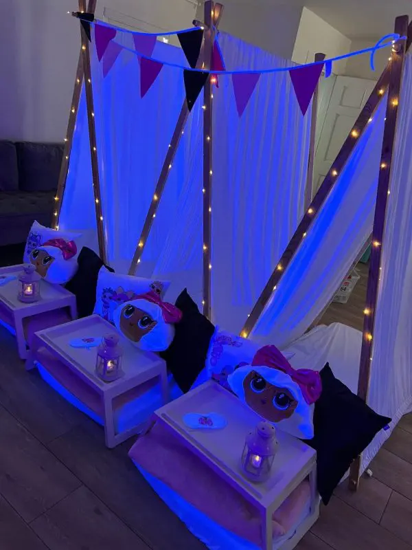 Indoor camping setup with party themes, including three beds under a white canopy, decorated with purple string lights and flags, each bed containing a plush toy and a serving tray.