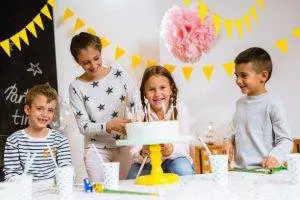 Four children smiling at a slumber party, with a girl about to blow out candles on a cake on a table decorated with confetti and party accessories.