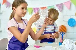 Girl in apron examines a paper snowflake while boy cuts paper at a decorated craft table for a slumber party.