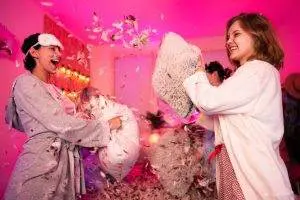 Two women having a pillow fight in a room lit by pink light, feathers flying around them during an Ultimate Canopy sleepover.