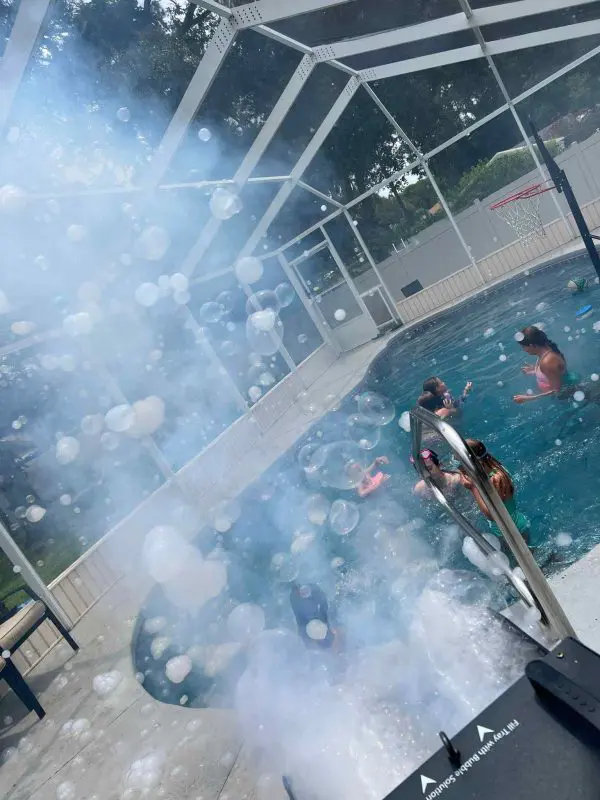 People enjoying a sunny day in a screened-in pool with a basketball hoop, a Frogger F4 Bubble Fogger Rental visible in the foreground.