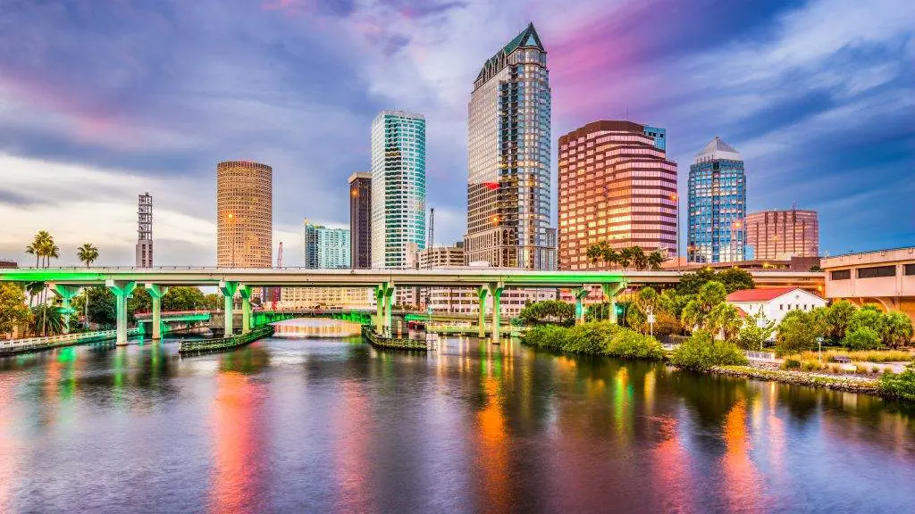 Panoramic view of a vibrant event tampa party rentals and a city skyline with a sunset, featuring modern skyscrapers and a green-lit bridge over a reflective river during a unique slumber party.