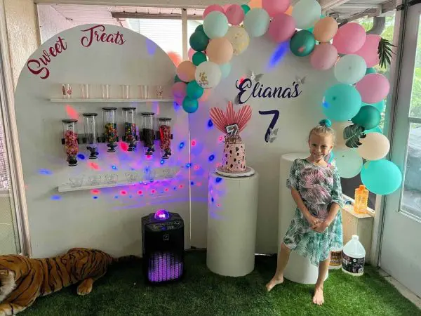 A young girl smiling at a colorful birthday party setup with balloons, a candy station, and a disco light, showcasing various party themes, with text "eliana's 7" on the backdrop.