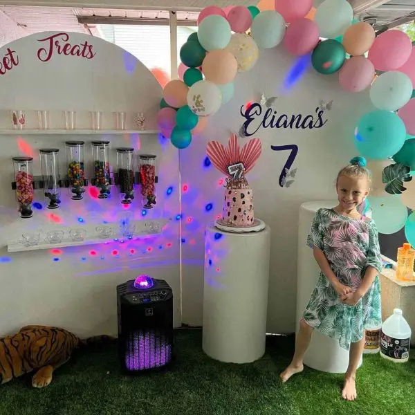 A young girl smiling at a colorful birthday party setup with balloons, a candy station, and a disco light, showcasing various party themes, with text "eliana's 7" on the backdrop.