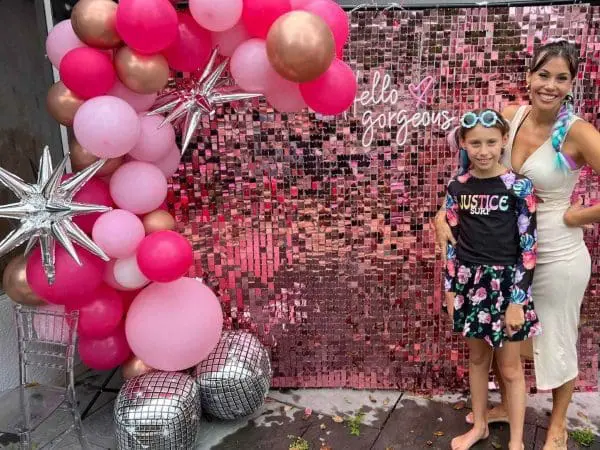 A woman and a young girl pose at a party by a pink backdrop with "hello gorgeous" sign, decorated with pink balloons and silver stars, perfect for event themes.