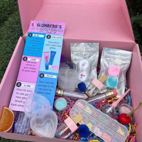 An open pink DIY Lip Gloss Party Box containing various tubes, glitter packets, mixing tools, and colorful decorations on a grass background.