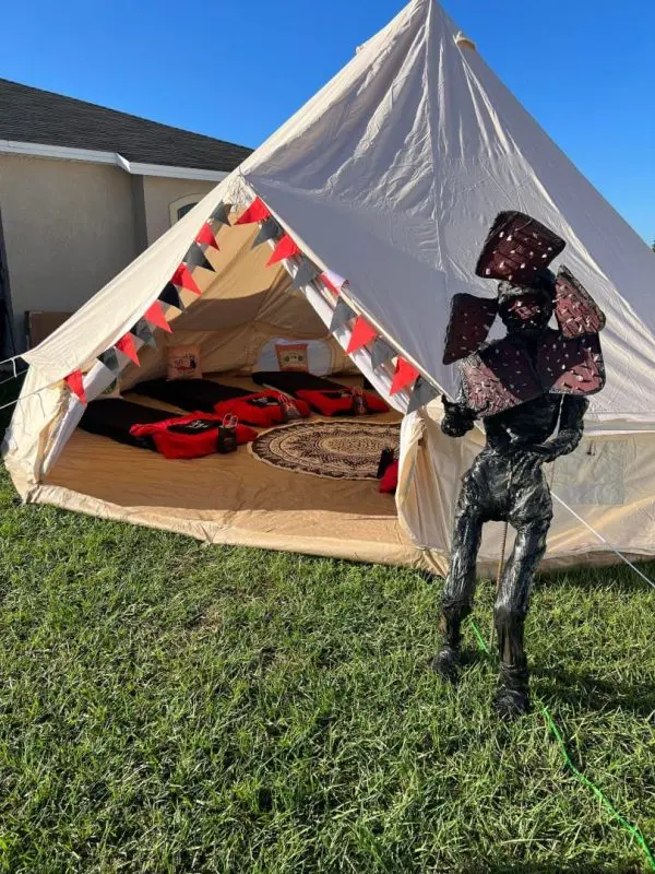 A person in a reflective polygonal suit stands in front of a large canvas tent decorated with bunting for outdoor glamping, set up on a grassy lawn.