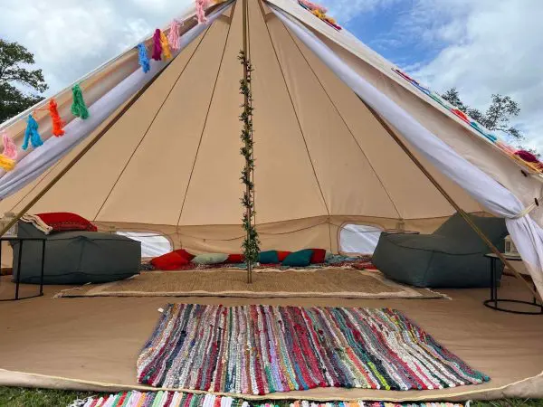 Large tent with open flap, decorated with colorful flags, furnished with bean bags, cushions, and a striped rug on a grassy Lakeland field.