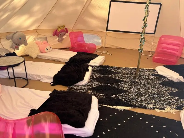 Interior of a tent with plush toys on beds, pink plastic chairs, a black rug, and scattered white petals, perfect for theme parties.